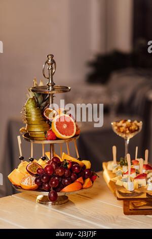 Ein Obstteller steht auf einem Holztisch, der für den Urlaub bedeckt ist.Tropisches Obstsortiment auf einem weißen Teller mit frischem Saft, textiler grauer Backgroun Stockfoto