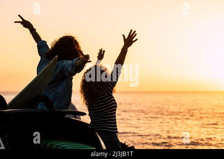 Reisen und Glücklichsein. Ein Paar Frauen, die die Arme gegen einen wunderschönen goldenen Sonnenuntergang auf dem Meer in der Nähe eines Autos erheben. Menschen in freundschaftlichsip Stockfoto