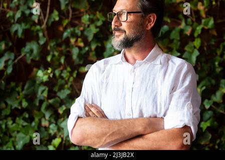Side Corporate Portrait von reifen bärtigen Mann mit Brillen lächeln mit gekreuzten Armen und grünen Blättern Garten Hintergrund. Selbstbewusster männlicher Erwachsener, Peopl Stockfoto