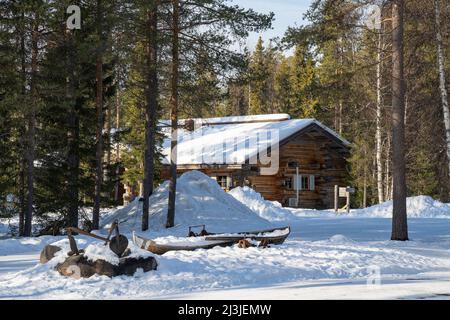 Rovaniemi, Finnland - 17.. März 2022: Eine Holzhütte und ein altes Boot in einem verschneiten Tannenwald an einem sonnigen Wintertag. Stockfoto