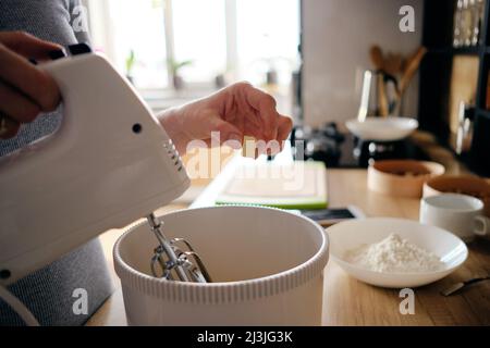 Eine Frau, die in der Küche mit einem Handmixer an der Theke steht, um frischeste Zutaten in einer weißen Rührschüssel zu schlagen, die auf dem Tisch liegen Stockfoto
