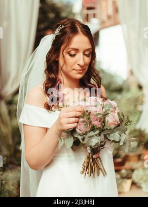 Braut in einem Kleid, das in einem grünen Garten steht und einen Hochzeitsstrauß aus Blumen und grünen Körnereffekten hält Stockfoto