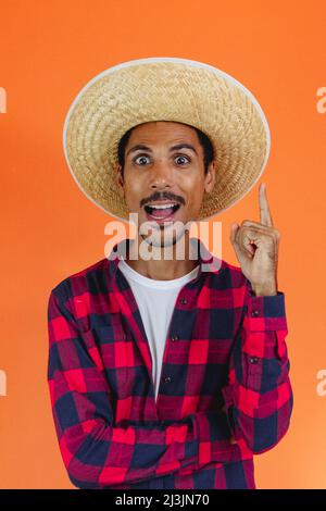 Schwarzer Mann mit Junina Party Outfit zeigt isoliert auf orangenen Hintergrund. Junger Mann in traditioneller Kleidung für Festa Junina - Brasilianischer Juni fe Stockfoto