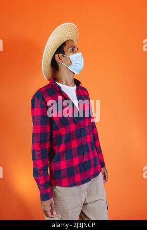 Schwarzer Mann mit Junina Party Outfit und Pandemiemaske isoliert auf orangefarbenem Hintergrund. Junger Mann in traditioneller Kleidung für Festa Junina - Brasilianer Stockfoto