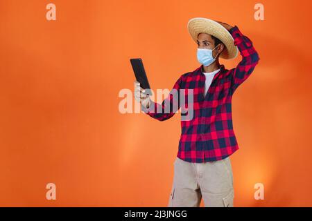 Schwarzer Mann mit Junina Party Outfit und Pandemiemaske isoliert auf orangefarbenem Hintergrund. Junger Mann in traditioneller Kleidung für Festa Junina - Brasilianer Stockfoto