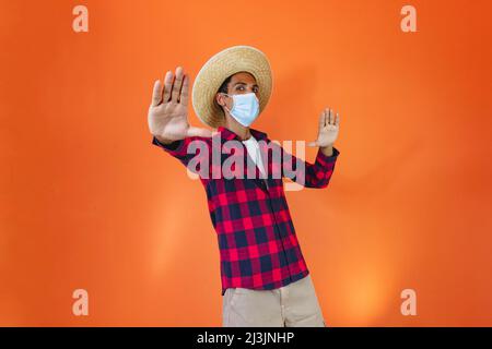 Schwarzer Mann mit Junina Party Outfit und Pandemiemaske isoliert auf orangefarbenem Hintergrund. Junger Mann in traditioneller Kleidung für Festa Junina - Brasilianer Stockfoto