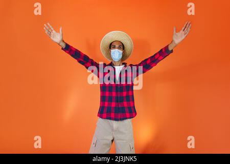 Schwarzer Mann mit Junina Party Outfit und Pandemiemaske isoliert auf orangefarbenem Hintergrund. Junger Mann in traditioneller Kleidung für Festa Junina - Brasilianer Stockfoto