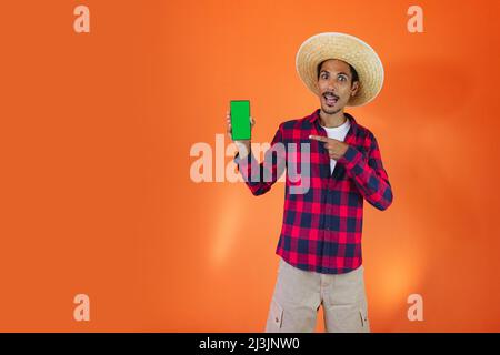 Schwarzer Mann mit Junina Party Outfit hält ein Tablet oder Handy isoliert auf orangefarbenem Hintergrund. Junger Mann in traditioneller Kleidung für Festa Junina - Stockfoto