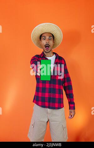 Schwarzer Mann mit Junina Party Outfit hält ein Tablet oder Handy isoliert auf orangefarbenem Hintergrund. Junger Mann in traditioneller Kleidung für Festa Junina - Stockfoto