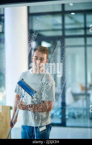 Saubere Fenster machen wirklich ein Büro. Aufnahme eines jungen Mannes, der die Fenster im Büro putzte. Stockfoto