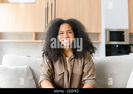 Porträt einer ruhigen und charmanten schönen dunkelhaarigen Frau. Die hübsche multirassische Dame in legerer Kleidung blickt auf die Kamera und lächelt, sie sitzt auf dem bequemen weißen Sofa Stockfoto