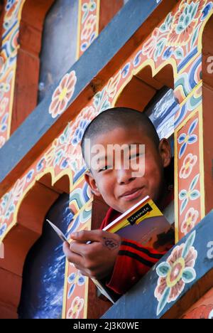 Im Kloster Gangteng Gompa in Phobjika, Bhutan, neigt sich ein junger Mönch durch ein Fenster, während er sein Schulbuch in der Hand hält: Phobjika, Bhutan Stockfoto