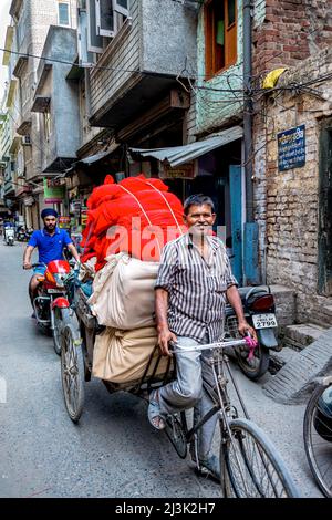 Ein Mann fährt mit dem Fahrrad und zieht einen beladenen Anhänger hinter sich, während ein Mann auf einem Motorrad auf einer Straße in Indien folgt; Amritsar, Punjab, Indien Stockfoto