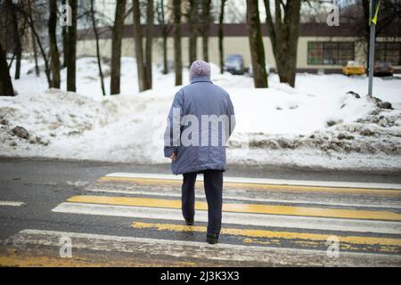 Frau überquert Straße auf Zebra. Rentner in Russland geht die Straße hinunter. Der Mensch geht auf Fußgängerüberwegen. Aufnahmen von hinten. Stockfoto
