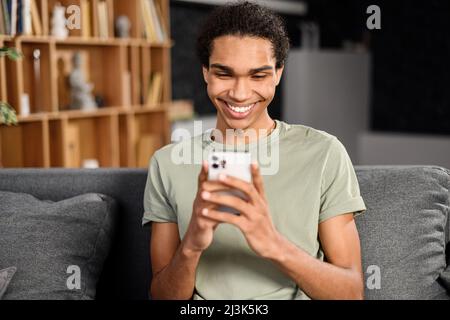 Hübscher Hipster-Typ im lässigen T-Shirt sitzt auf einem bequemen Sofa und verbringt Freizeit online am Telefon, fröhlicher multirassischer Teenager-Typ ruht sich aus, scrollt in den Neuigkeiten, schaut auf den Bildschirm des Geräts, lacht Stockfoto