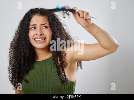 Ich wünschte, es gäbe eine einfachere Möglichkeit, meine Haare zu entwirren. Aufnahme einer Frau, die runzelte, während sie ihr Haar vor einem grauen Hintergrund kämmte. Stockfoto