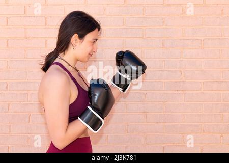 Eine junge kaukasische Frau mit schwarzen Boxhandschuhen in einer defensiven Position von der Seite aus gesehen. Im Hintergrund ist eine Ziegelmauer. Stockfoto