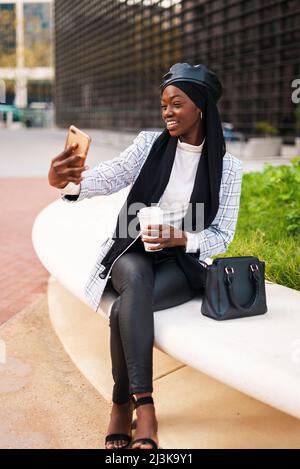 Lächelnde afroamerikanische Frau in eleganter Freizeitkleidung und muslimischem Kopftuch, die mit Kaffee zum Mitnehmen sitzt und in der Stadt Selbstporträts auf dem Handy aufführt Stockfoto