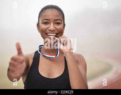 Der Erfolg schmeckt sicher süß. Aufnahme einer jungen Athletin, die den Daumen hoch gab, während sie ihre Medaille beißt. Stockfoto