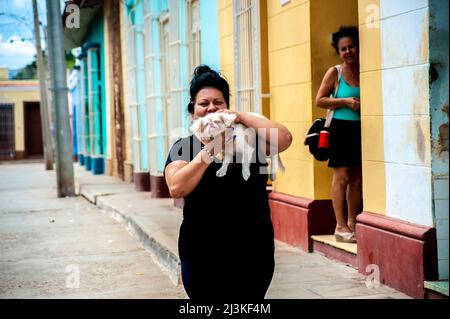 Frau hält und küsst ihren Hund auf einer Straße in Trinidad, Kuba, während ihre Freundin schaut, lächelt und lacht. Stockfoto