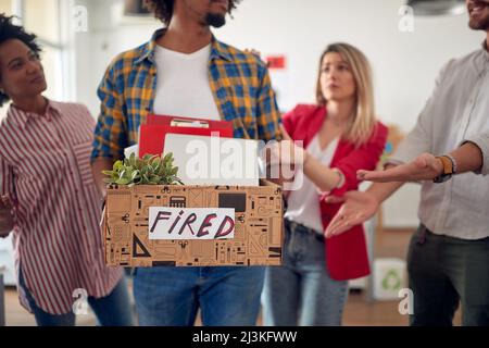 Kollegen trösten den traurigen jungen männlichen Mitarbeiter, als er das Büro verlässt, nachdem er von einem Job entlassen wurde, den er liebt. Mitarbeiter, Job, Büro Stockfoto