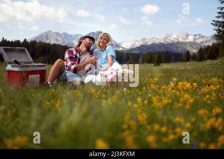 Attraktives junges Paar mit Picknick in der Natur. Mann, der einer glücklichen Frau eine Blume aus dem Dandelion gibt. Spaß, Zweisamkeit, Naturkonzept. Stockfoto