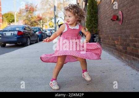 Das junge Mädchen steht auf einem Bürgersteig in der Stadt, der in ihrem rot-weißen Kleid in den Kindergingham-Farben wirbelt; Toronto, Ontario, Kanada Stockfoto