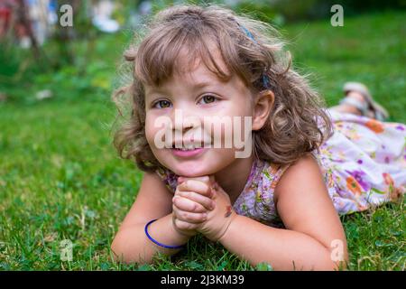 Porträt eines Vorschulmädchen mit blondem lockigen Haar und braunen Augen, das auf dem Gras liegt und die Hände vor ihr gefaltet sind; Toronto, Ontario, Kanada Stockfoto