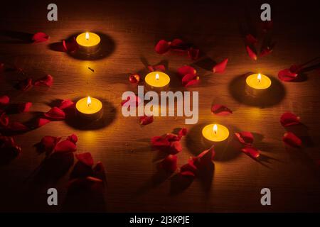 Beleuchtete Teelichtkerzen und rote Rosenblätter, die auf einer Oberfläche verstreut sind, schaffen eine romantische Umgebung; Studio Stockfoto