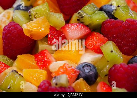 Farbenfroher Obstsalat in einer Schüssel auf schwarzem Hintergrund. Nahaufnahme von frischem und schmackhaftem Salat mit Orange, Apfel, Beeren, Granatapfel, Kiwi, ananas Stockfoto