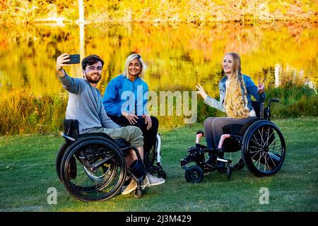 Junge querschnittsgelähmte Männer und Frauen im Rollstuhl, die an einem schönen Herbsttag mit einem Smartphone in einem Park ein Selbstporträt machen Stockfoto