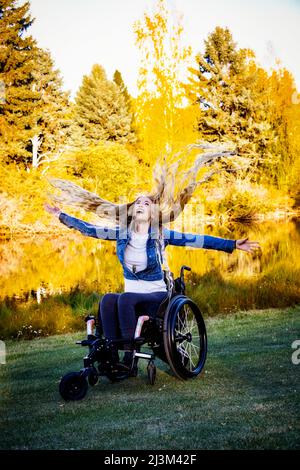 Junge querschnittsgelähmte Frau in ihrem Rollstuhl in einem Park an einem schönen Herbsttag, sich sorglos fühlend mit ausgestreckten Armen und ihren langen Haaren ... Stockfoto