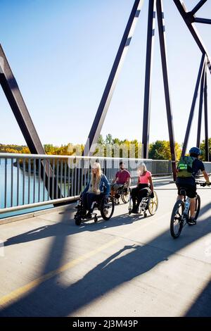 Drei junge querschnittsgelähmende Freunde verbringen an einem schönen Tag Zeit miteinander und bewegen sich über eine Parkbrücke in ihren Rollstühlen in einem Stadtpark Stockfoto