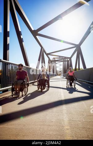 Drei junge querschnittsgelähmende Freunde verbringen an einem schönen Tag Zeit miteinander und bewegen sich über eine Parkbrücke in ihren Rollstühlen in einem Stadtpark Stockfoto