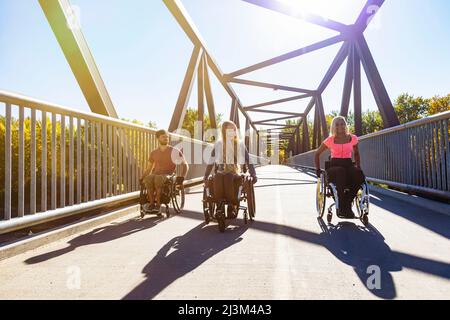 Drei junge querschnittsgelähmende Freunde verbringen an einem schönen Tag Zeit miteinander und bewegen sich über eine Parkbrücke in ihren Rollstühlen in einem Stadtpark Stockfoto