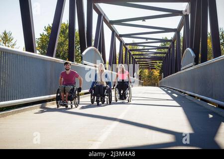 Drei junge querschnittsgelähmende Freunde verbringen an einem schönen Tag Zeit miteinander und bewegen sich über eine Parkbrücke in ihren Rollstühlen in einem Stadtpark Stockfoto