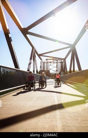 Drei junge querschnittsgelähmende Freunde verbringen an einem schönen Tag Zeit miteinander und bewegen sich über eine Parkbrücke in ihren Rollstühlen in einem Stadtpark Stockfoto