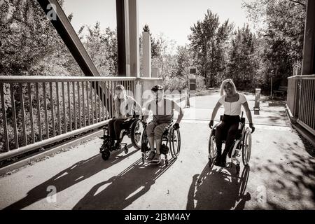 Drei junge querschnittsgelähmende Freunde verbringen an einem schönen Tag Zeit miteinander und bewegen sich über eine Parkbrücke in ihren Rollstühlen in einem Stadtpark Stockfoto