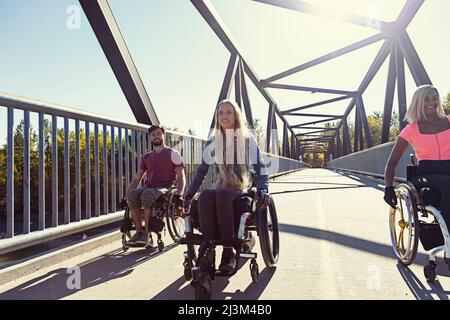 Drei junge querschnittsgelähmende Freunde verbringen an einem schönen Tag Zeit miteinander und bewegen sich über eine Parkbrücke in ihren Rollstühlen in einem Stadtpark Stockfoto