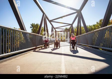 Drei junge querschnittsgelähmende Freunde verbringen an einem schönen Tag Zeit miteinander und bewegen sich über eine Parkbrücke in ihren Rollstühlen in einem Stadtpark Stockfoto