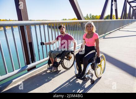 Zwei junge querschnittsgelähmende Freunde verbringen an einem schönen Tag Zeit miteinander und bewegen sich über eine Parkbrücke in ihren Rollstühlen in einem Stadtpark Stockfoto