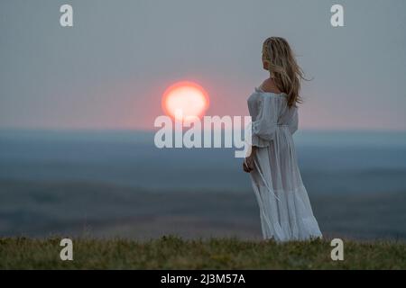 Frau, die in einem weißen Kleid auf einem Bluff steht und dabei den Sonnenuntergang beobachtet; Saskatchewan, Kanada Stockfoto