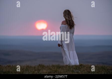 Frau, die in einem weißen Kleid auf einem Bluff steht und dabei den Sonnenuntergang beobachtet; Saskatchewan, Kanada Stockfoto