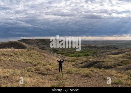 Frau, die mit erhobenen Armen steht, während sie eine wunderschöne aussicht im Grassands National Park, Val Marie, Saskatchewan, Kanada, betrachtet Stockfoto
