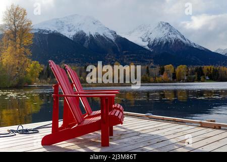 Zwei rote Stühle auf einer Terrasse auf dem ruhigen See Kathlyn, umgeben von herbstbunten Bäumen und schneebedeckten Coast Mountains, vom Lakedrop Inn, Wa... Stockfoto