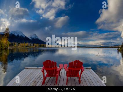 Zwei rote Stühle auf einer Terrasse auf dem ruhigen See Kathlyn, umgeben von herbstbunten Bäumen und schneebedeckten Coast Mountains, vom Lakedrop Inn, Wa... Stockfoto