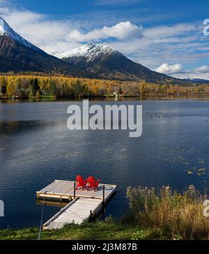 Zwei rote Stühle auf einer Terrasse auf dem ruhigen See Kathlyn, umgeben von herbstbunten Bäumen und schneebedeckten Coast Mountains, vom Lakedrop Inn, Wa... Stockfoto