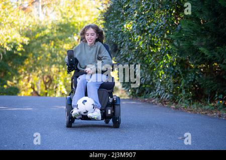 Das elfjährige Mädchen mit Ullrich kongenitaler Muskeldystrophie spielt in ihrem Rollstuhl in der Nähe ihres Heims Fußball Stockfoto