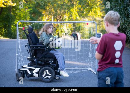 Das elfjährige Mädchen mit Ullrich kongenitaler Muskeldystrophie spielt mit einem Freund in der Nähe ihres Heims im Rollstuhl Fußball Stockfoto