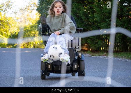 Das elfjährige Mädchen mit Ullrich kongenitaler Muskeldystrophie spielt in ihrem Rollstuhl in der Nähe ihres Heims Fußball Stockfoto